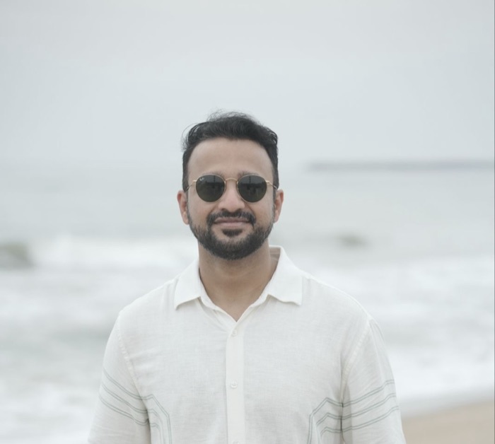 Sethu Prabakaran on a beach in a white linen shirt and round sunglasses